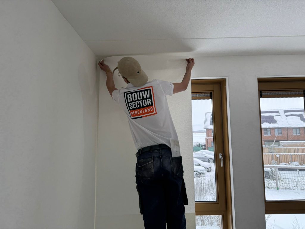 Behanger van Bouwsector Nederland trekt een baan behang strak tot tegen het plafond naast een raam met houten kozijn