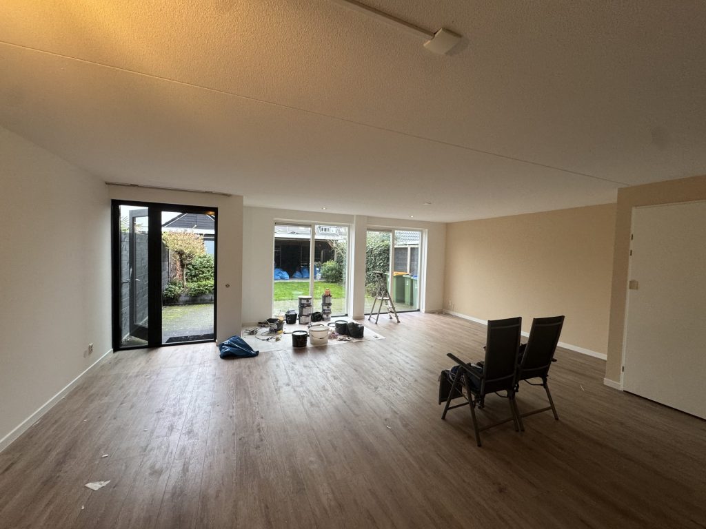Ruime woonkamer van een bestaand huis met lichte wanden, een houten vloer en tuindeuren, tijdens het werk