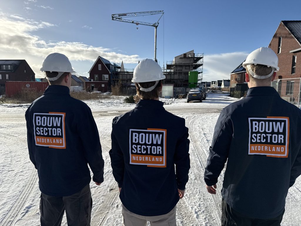 Drie vakmensen van Bouwsector Nederland met witte helm en logo op de jas, bij huizen in aanbouw en een bouwkraan